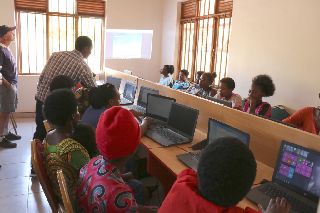 Children sat in classroom in front of computers while teacher explains something.